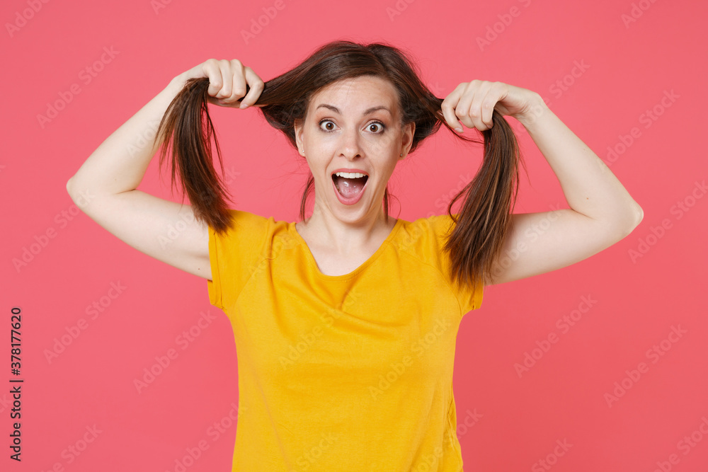 Excited surprised shocked young brunette woman 20s in yellow casual t-shirt posing holding hair like ponytails fooling around keeping mouth open isolated on pink color wall background studio portrait.