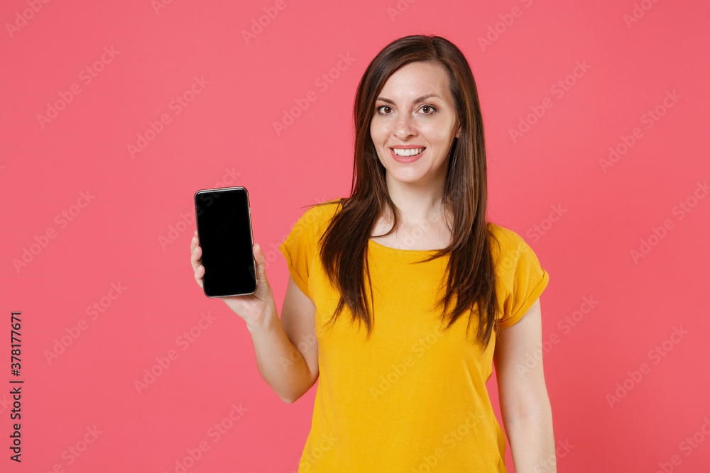 Smiling young brunette woman 20s wearing yellow casual t-shirt posing hold mobile phone with blank empty screen mock up copy space looking camera isolated on pink color background studio portrait. Smiling young brunette woman 20s wearing yellow casual t-shirt posing hold mobile phone with blank empty screen mock up copy space looking camera isolated on pink color background studio portrait.