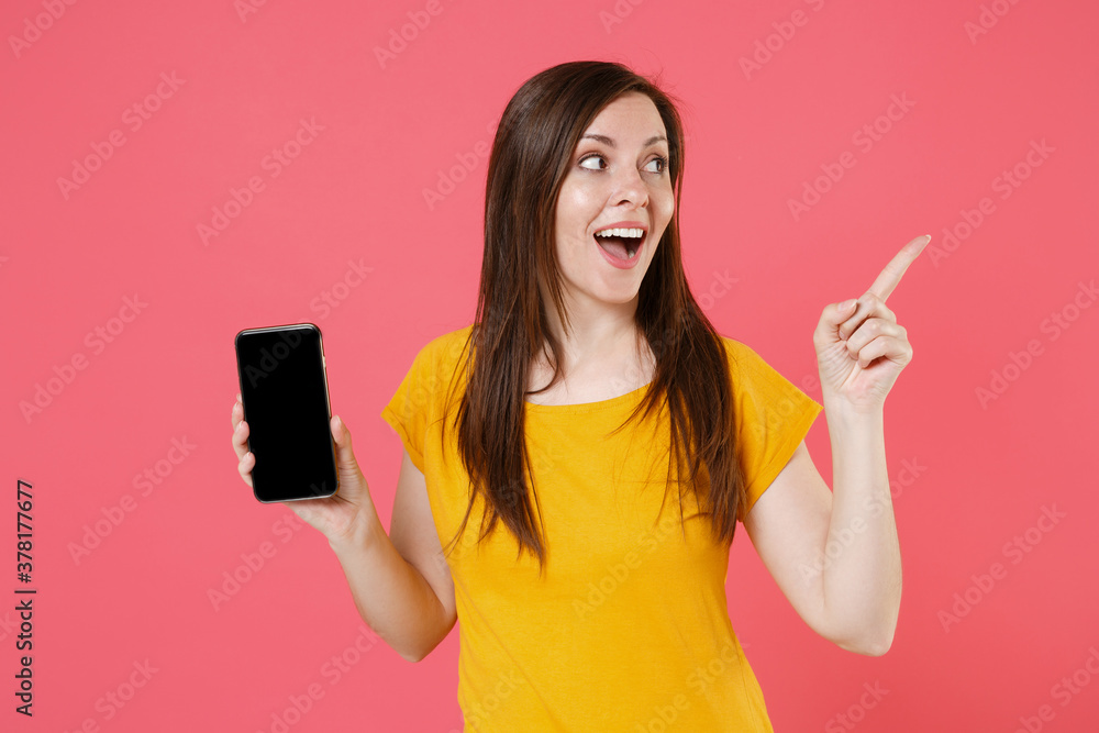 Excited young brunette woman in yellow casual t-shirt posing hold mobile phone with blank empty screen mock up copy space pointing index finger aside isolated on pink color background studio portrait. Excited young brunette woman in yellow casual t-shirt posing hold mobile phone with blank empty screen mock up copy space pointing index finger aside isolated on pink color background studio portrait.