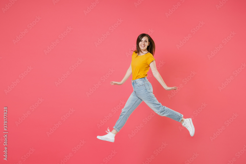 Full length side view portrait of smiling young brunette woman 20s wearing yellow casual t-shirt posing jumping spreading hands and legs looking camera isolated on pink color wall background studio.