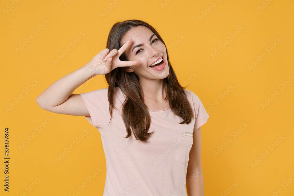 Cheerful funny pretty young brunette woman 20s wearing pastel pink casual t-shirt standing posing showing victory sign looking camera isolated on bright yellow color wall background studio portrait.