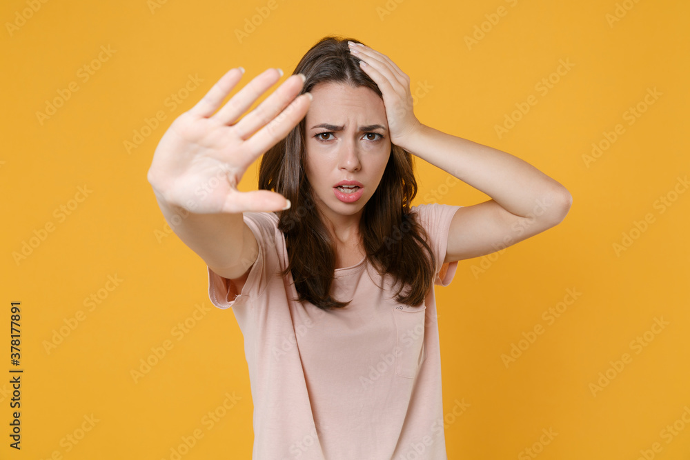 Worried shocked young brunette woman wearing pastel pink casual t-shirt stand posing showing stop gesture with palm put hand on head looking camera isolated on yellow color background studio portrait. Worried shocked young brunette woman wearing pastel pink casual t-shirt stand posing showing stop gesture with palm put hand on head looking camera isolated on yellow color background studio portrait.