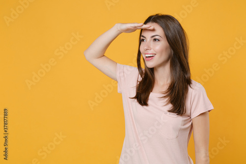 Smiling funny young brunette woman 20s wearing pastel pink casual t-shirt standing posing holding hand at forehead looking far away distance isolated on bright yellow color background studio portrait.