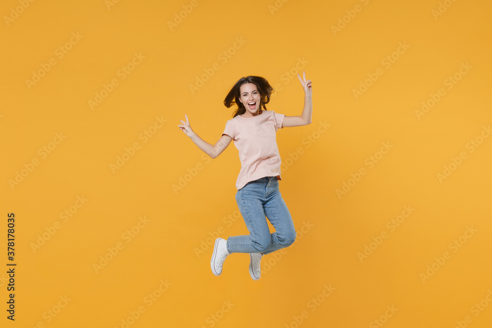 Full length portrait of cheerful funny young woman 20s wearing pastel pink casual t-shirt posing jumping showing victory sign looking camera isolated on bright yellow color wall background studio. Full length portrait of cheerful funny young woman 20s wearing pastel pink casual t-shirt posing jumping showing victory sign looking camera isolated on bright yellow color wall background studio.