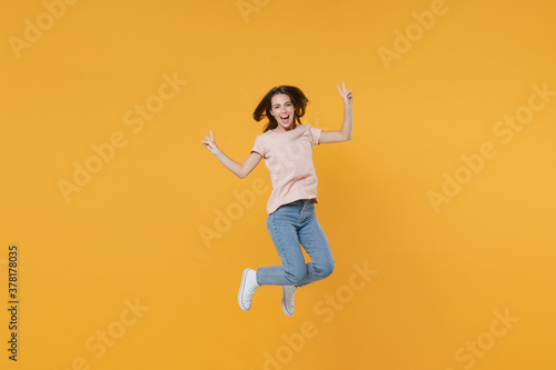 Full length portrait of cheerful funny young woman 20s wearing pastel pink casual t-shirt posing jumping showing victory sign looking camera isolated on bright yellow color wall background studio.