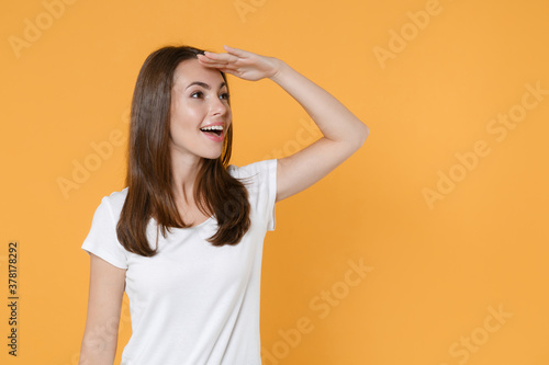 Excited young brunette woman 20s in white blank empty design casual t-shirt posing standing holding hand at forehead looking far away distance isolated on yellow color wall background studio portrait.