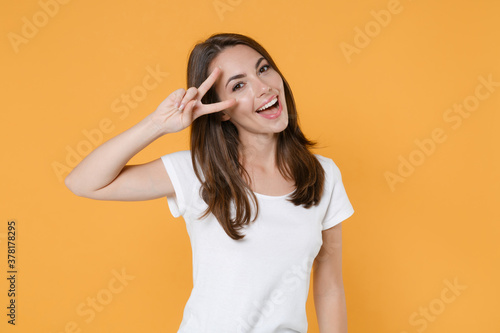 Beautiful funny cheerful pretty young brunette woman 20s wearing white blank empty design casual t-shirt posing standing showing victory sign isolated on yellow color wall background studio portrait.