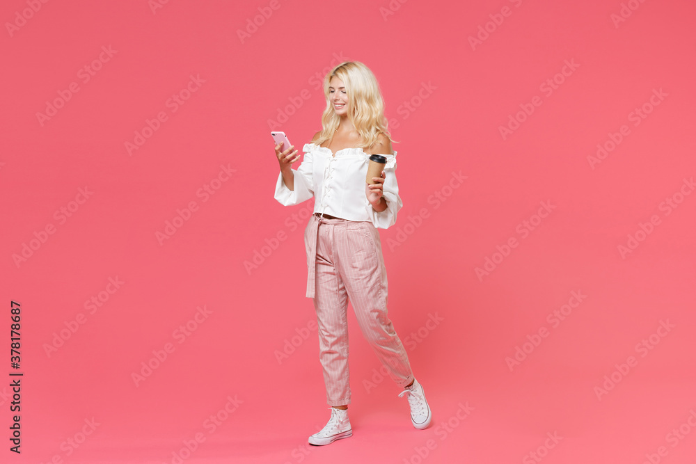 Full length portrait of smiling young blonde woman in white casual clothes using mobile cell phone typing sms message hold paper cup of coffee or tea isolated on bright pink colour background studio. Full length portrait of smiling young blonde woman in white casual clothes using mobile cell phone typing sms message hold paper cup of coffee or tea isolated on bright pink colour background studio.