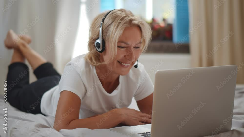 Cheerful grandmother in headset talking with relatives via laptop lying on bed Cheerful grandmother in headset talking with relatives via laptop lying on bed