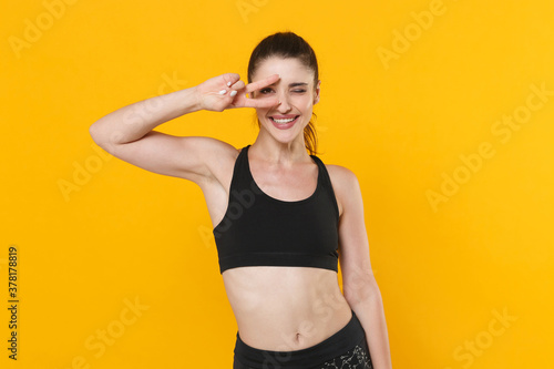 Fototapeta Naklejka Na Ścianę i Meble -  Smiling blinking young fitness sporty woman 20s wearing black sportswear posing training working out showing victory sign looking camera isolated on bright yellow color background studio portrait.