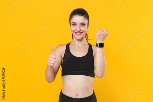 Smiling young fitness sporty woman 20s wearing black sportswear posing working out training wearing smart watch on hand showing thumb up isolated on bright yellow color background studio portrait.
