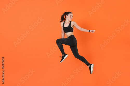 Full length side view of portrait of funny young fitness sporty woman 20s in black sportswear posing training working out jumping like running looking aside isolated on orange color background studio.