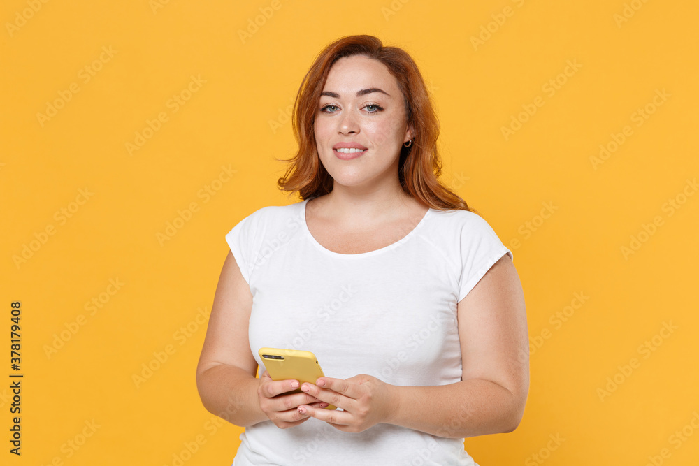 Smiling young redhead plus size body positive female woman girl 20s in white casual t-shirt posing using mobile cell phone, typing sms message isolated on yellow color wall background studio portrait. Smiling young redhead plus size body positive female woman girl 20s in white casual t-shirt posing using mobile cell phone, typing sms message isolated on yellow color wall background studio portrait.