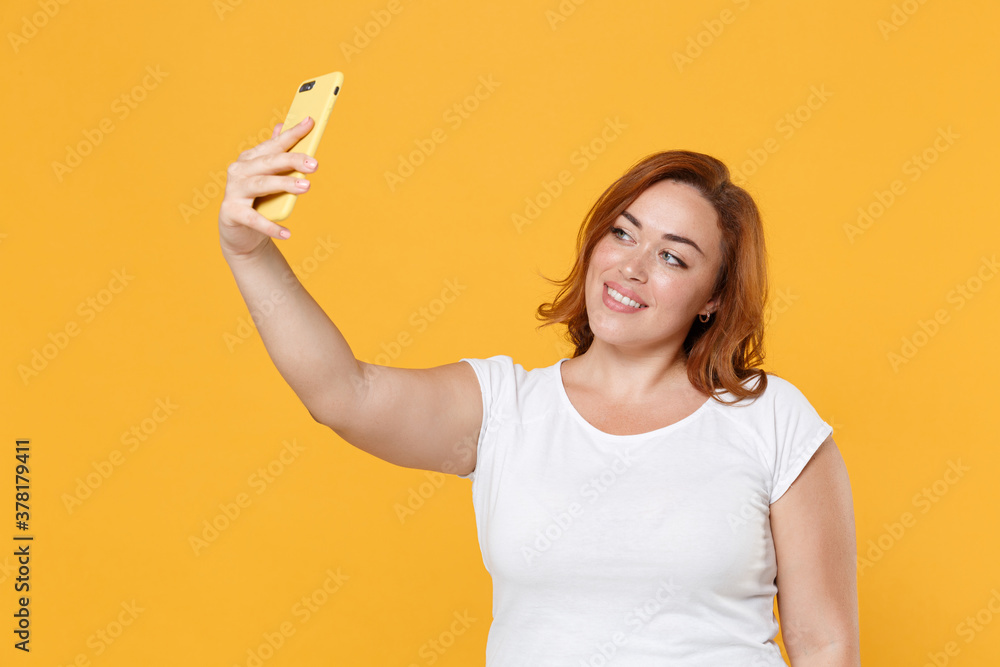 Smiling beautiful young redhead plus size body positive female woman girl 20s in white casual t-shirt posing doing selfie shot on mobile phone isolated on yellow color wall background studio portrait. Smiling beautiful young redhead plus size body positive female woman girl 20s in white casual t-shirt posing doing selfie shot on mobile phone isolated on yellow color wall background studio portrait.