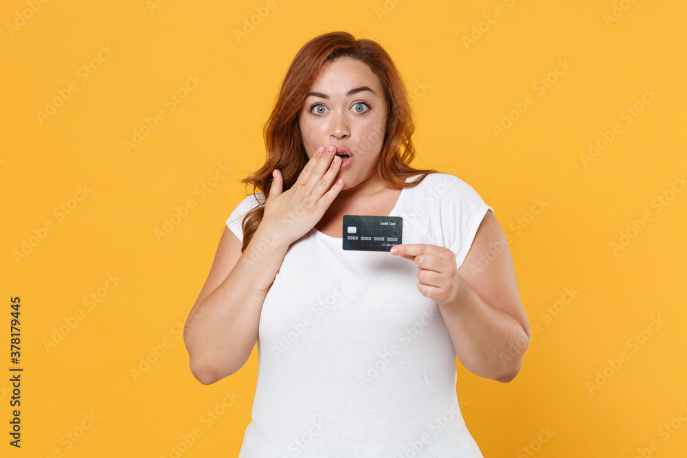 Shocked young redhead plus size body positive female woman girl 20s in white casual t-shirt posing hold credit bank card covering mouth with hand isolated on yellow color background studio portrait. Shocked young redhead plus size body positive female woman girl 20s in white casual t-shirt posing hold credit bank card covering mouth with hand isolated on yellow color background studio portrait.
