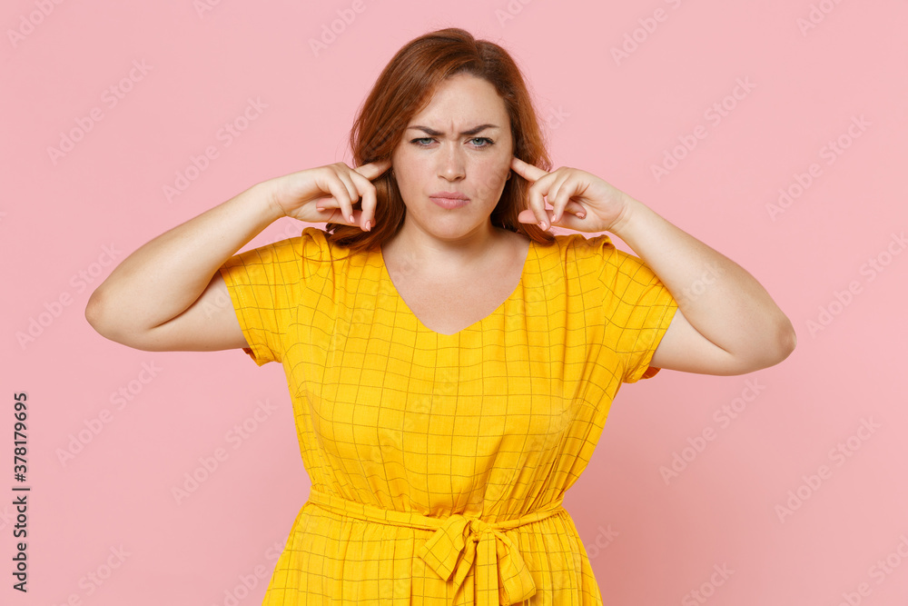 Dissatisfied concerned young redhead plus size body positive female woman girl 20s in yellow dress covering ears with fingers looking camera isolated on pastel pink color background studio portrait.