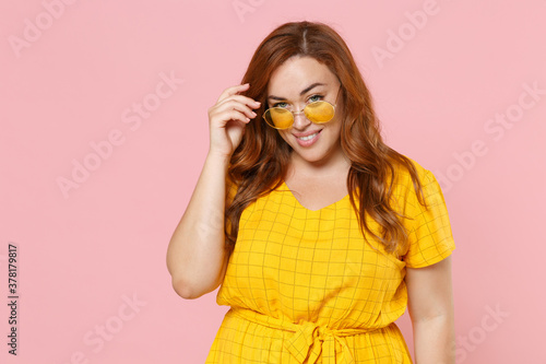 Smiling stunning beautiful young redhead plus size body positive female woman girl 20s in yellow dress eyeglasses posing put hand on head isolated on pastel pink color wall background studio portrait. © ViDi Studio