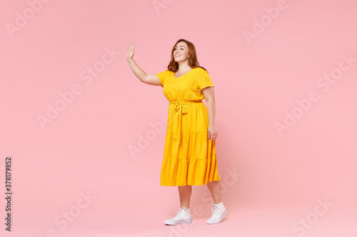 Full length portrait of smiling young redhead plus size body positive female woman 20s in yellow dress waving and greeting with hand as notices someone isolated on pastel pink color background studio.