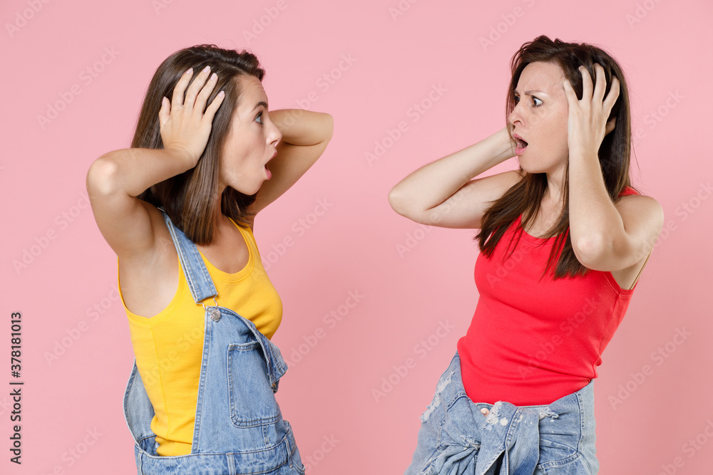 Two worried shocked puzzled young women friends 20s in casual denim clothes put hands on head keeping mouth open looking at each other isolated on pastel pink colour background, studio portrait. Two worried shocked puzzled young women friends 20s in casual denim clothes put hands on head keeping mouth open looking at each other isolated on pastel pink colour background, studio portrait.
