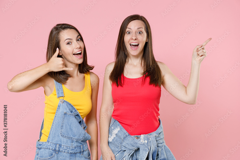 Two surprised young women friends in denim clothes doing phone gesture says call me back pointing index finger aside up on mock up copy space isolated on pastel pink colour background studio portrait. Two surprised young women friends in denim clothes doing phone gesture says call me back pointing index finger aside up on mock up copy space isolated on pastel pink colour background studio portrait.