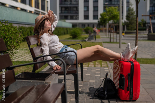 A beautiful young woman is sitting on a bench and put her legs on a red suitcase ouydoors photo