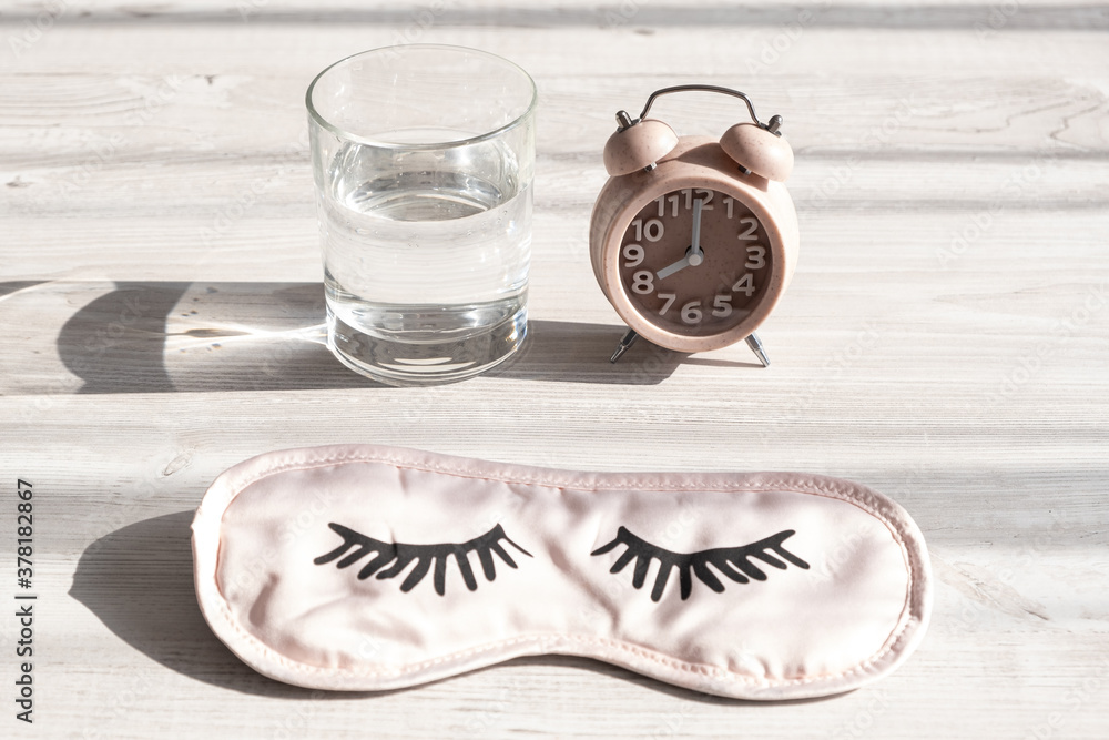 alarm clock and sleep mask, glass with clean water on a wooden white table in the morning sunlight. The concept of starting a new day, good morning or ending the day, late evening. Day planning alarm clock and sleep mask, glass with clean water on a wooden white table in the morning sunlight. The concept of starting a new day, good morning or ending the day, late evening. Day planning