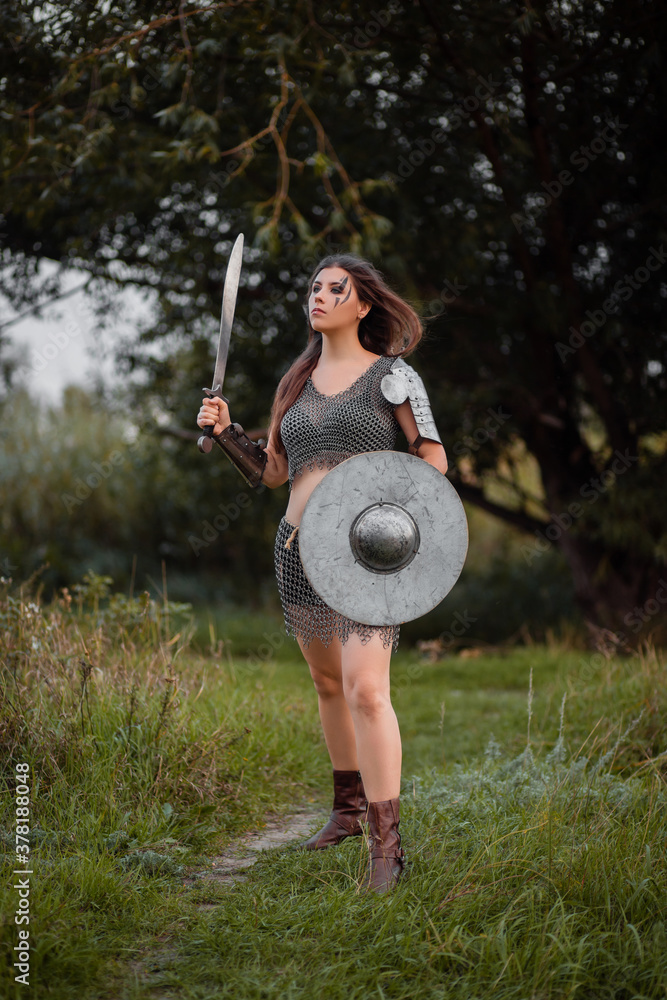 A medieval female warrior dressed in chain mail with a sword and shield in  her hands poses against the background of a forest. Fantasy costume, combat  makeup. Stock Photo