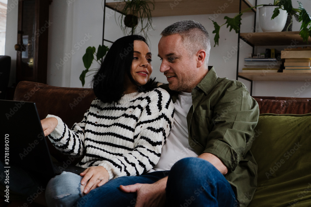 Romantic couple smiling at each other while waiting to chat with friends on video call using laptop sitting on sofa in lounge. Romantic couple smiling at each other while waiting to chat with friends on video call using laptop sitting on sofa in lounge.