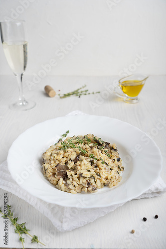 Creamy risotto or Italian arborio rice dish containing broth, mushrooms, thyme, olive oil and parmesan cheese served in plate with glass of white on white wooden background. Vertical orientation