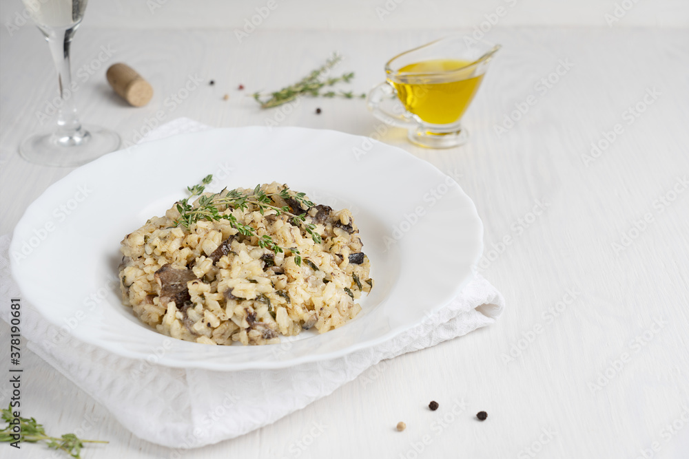 Creamy risotto or Italian arborio rice dish with mushrooms, thyme, olive oil and parmesan cheese served in plate with glass of white wine on towel on white wooden background. Image with copy space