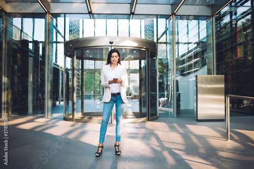 Beautiful 30s female in trendy smart casual wear standing near business center building checking notification on mobile phone, confident businesswoman reading message and chatting on cellular