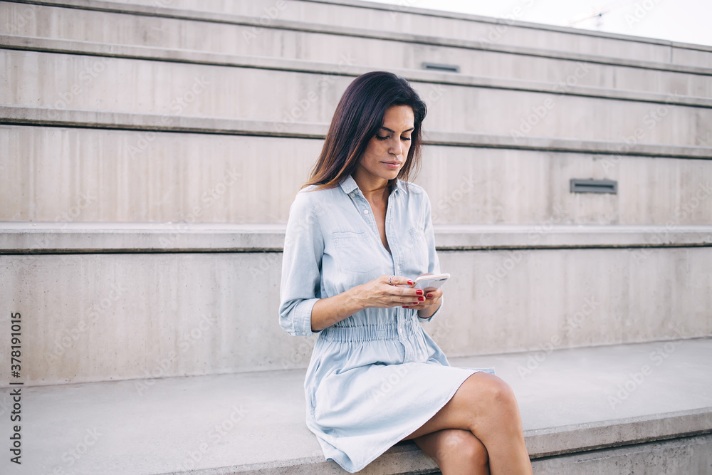 Serious caucasian 30s woman millennial sending text messages and reading information on mobile phone sitting outdoors, pensive female watching video online on smartphone resting on urban setting Serious caucasian 30s woman millennial sending text messages and reading information on mobile phone sitting outdoors, pensive female watching video online on smartphone resting on urban setting