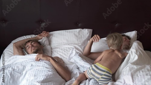 Morning in loving family. Cute child boy comes in bedroom to sleeping parents and start wakes them up. Mother and father sleeping together in bed at home, active little boy interrupts them.