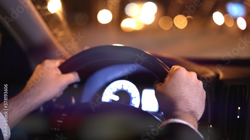 Male hands on steering wheel, backseat shot with view on road at night. Hands on wheel, man in black suit. Road with bokeh lights in dark night photo