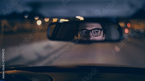 Driver's face is reflected in a rear view mirror, night shot, violet color, bokeh light. Tired concentrated driver in eyeglasses is reflected in a mirror in car, night road shot driving all night