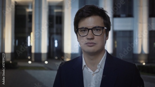 Serious businessman in eyeglasses and black suit at night outside, on background of blue buildings. Night city shot of man, looking straight at the camera