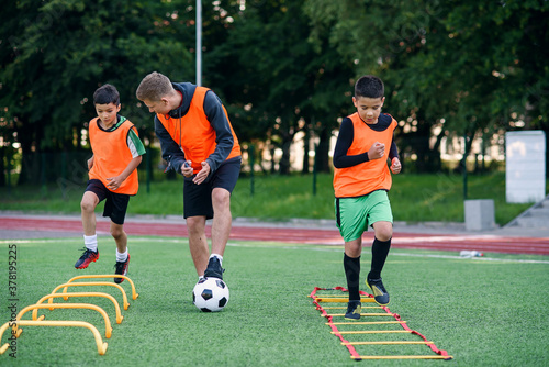 Confident soccer coach watches to his pupils making run exercises with overcoming obstacles with racks on soccer field