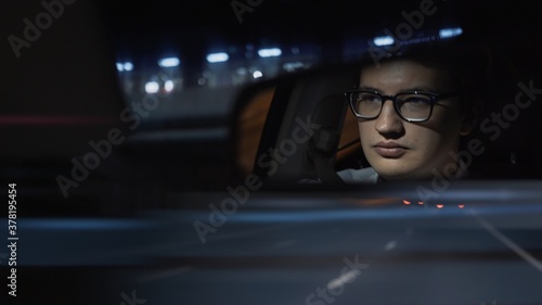Driver's face is reflected in a rear view mirror, night shot, dark blue road. Driver in eyeglasses is reflected in a mirror in car, night road shot driving all night