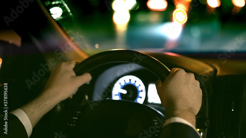 Male hands on steering wheel, backseat shot with view on road at night. Hands on wheel, man in black suit. Road with bokeh lights in dark night photo