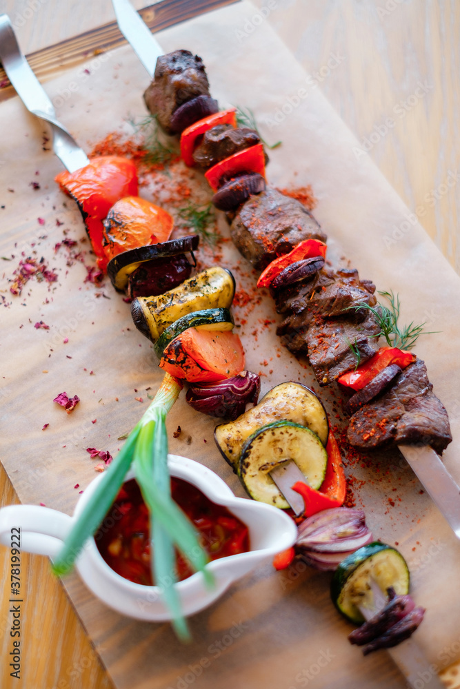Fototapeta premium Fried meat on a skewer with potatoes and vegetables. Grilled BBQ decorated with vegetables and sauce on wooden table and parchment paper.