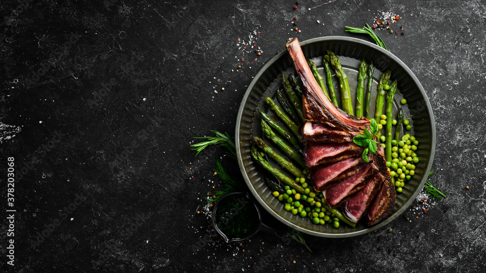 Tomahawk steak on the bone. Grilled steak with asparagus and green peas. On a black background. Top view. Free copy space. Tomahawk steak on the bone. Grilled steak with asparagus and green peas. On a black background. Top view. Free copy space.