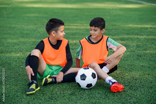 Good-looking joyful teen football players resting on the artificial grass covering of soccer field after training.