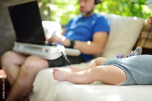 Young dad works remotely from home office with baby. Freelancer man holding his infant while using laptop. Workplace in living room. Working during quarantine.