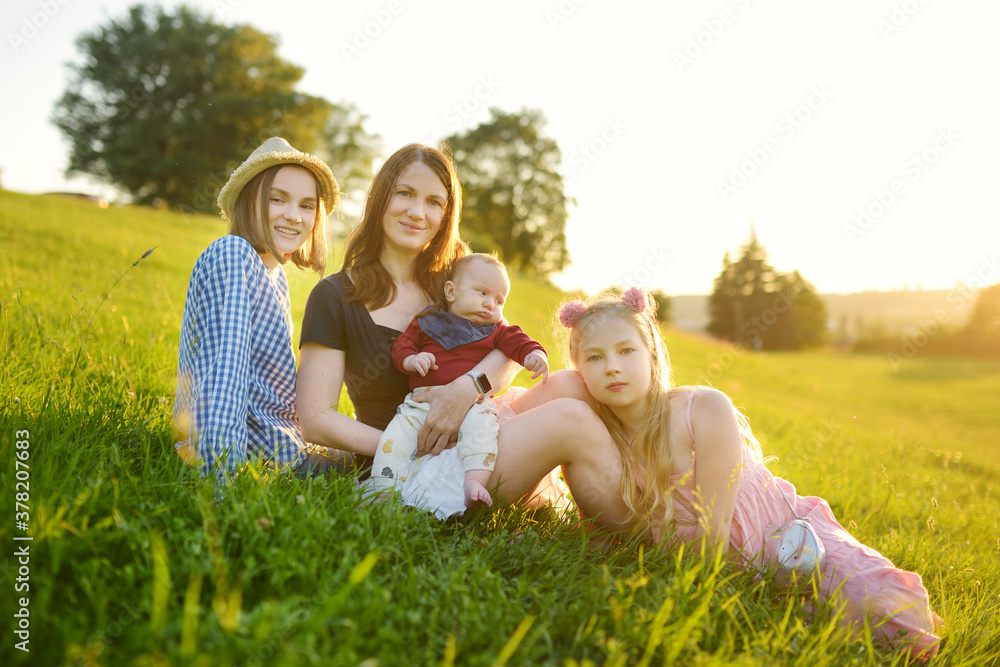 Fototapeta premium Mother and three children having fun on summer day in city park. Adorable baby boy being held by his mommy. Two older sisters hugging their mom and baby brother. Kids with large age gap.