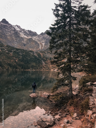 a young man a guy in a hood stands on the Big Stone in the middle of a clear azure green lake surrounded by high mountains and trees. Snow-covered tops of mountains and dry conifers in the fog. © Andrii