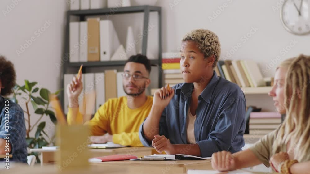 Group of young multiethnic students sitting at desks in classroom and raising arms, then young mixed-race woman asking question and having discussion with professor during college class Group of young multiethnic students sitting at desks in classroom and raising arms, then young mixed-race woman asking question and having discussion with professor during college class