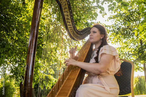 young woman in pink dress plays fairy music on a harp in the forest. Harmony with nature. Musical art. Classical concert
