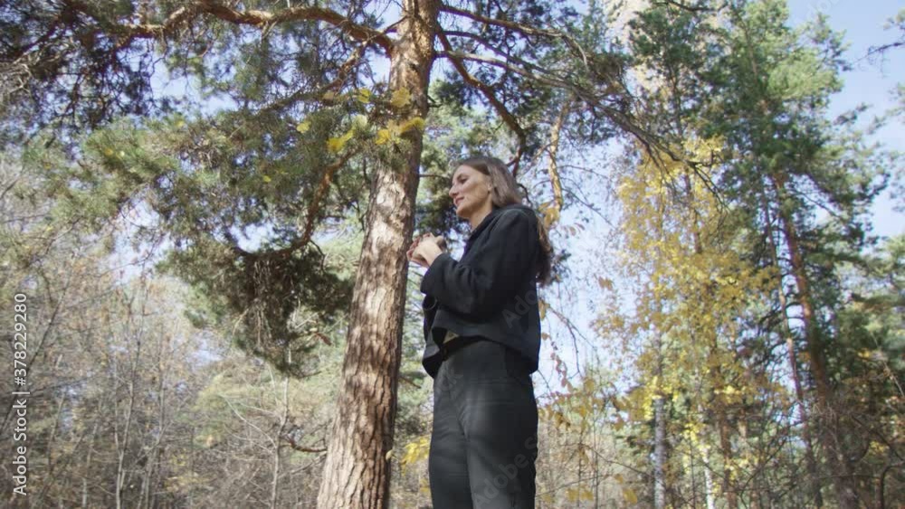 Dark-haired woman dressed in black sportswear warming up wrists while standing among trees in autumn forest. Low angle 4K 360 degree tracking arc shot. Dark-haired woman dressed in black sportswear warming up wrists while standing among trees in autumn forest. Low angle 4K 360 degree tracking arc shot.