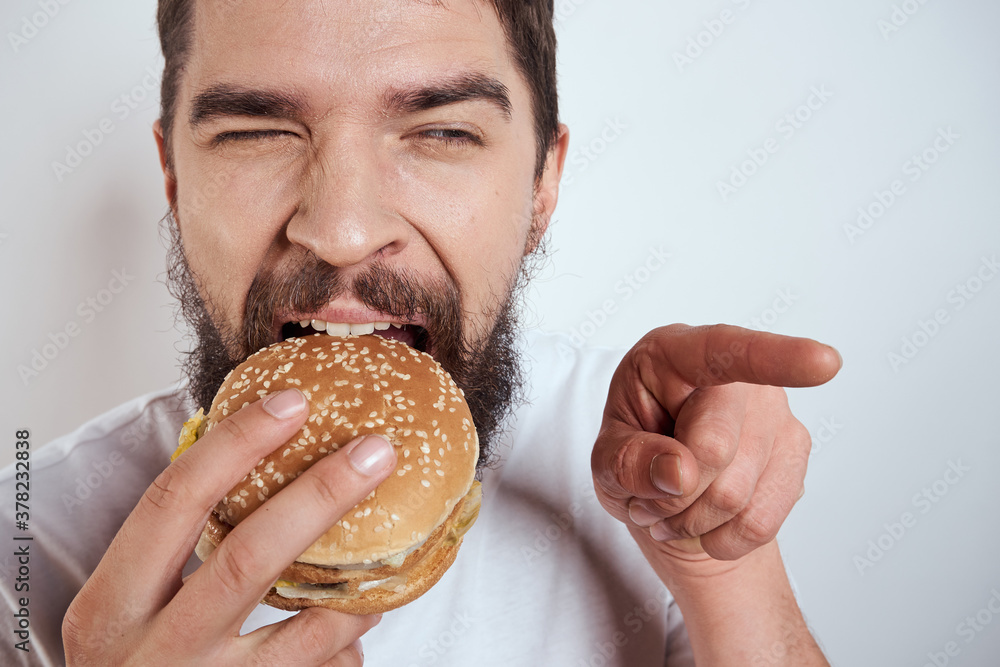 A man eating a hamburger on a light background in a white T-shirt cropped view close-up hunger fast food A man eating a hamburger on a light background in a white T-shirt cropped view close-up hunger fast food