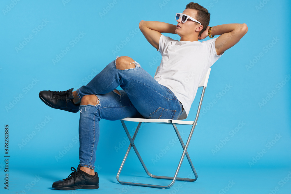 Relaxed guy in jeans with torn knees sits on a chair indoors on a blue background with full-length glasses on his face
